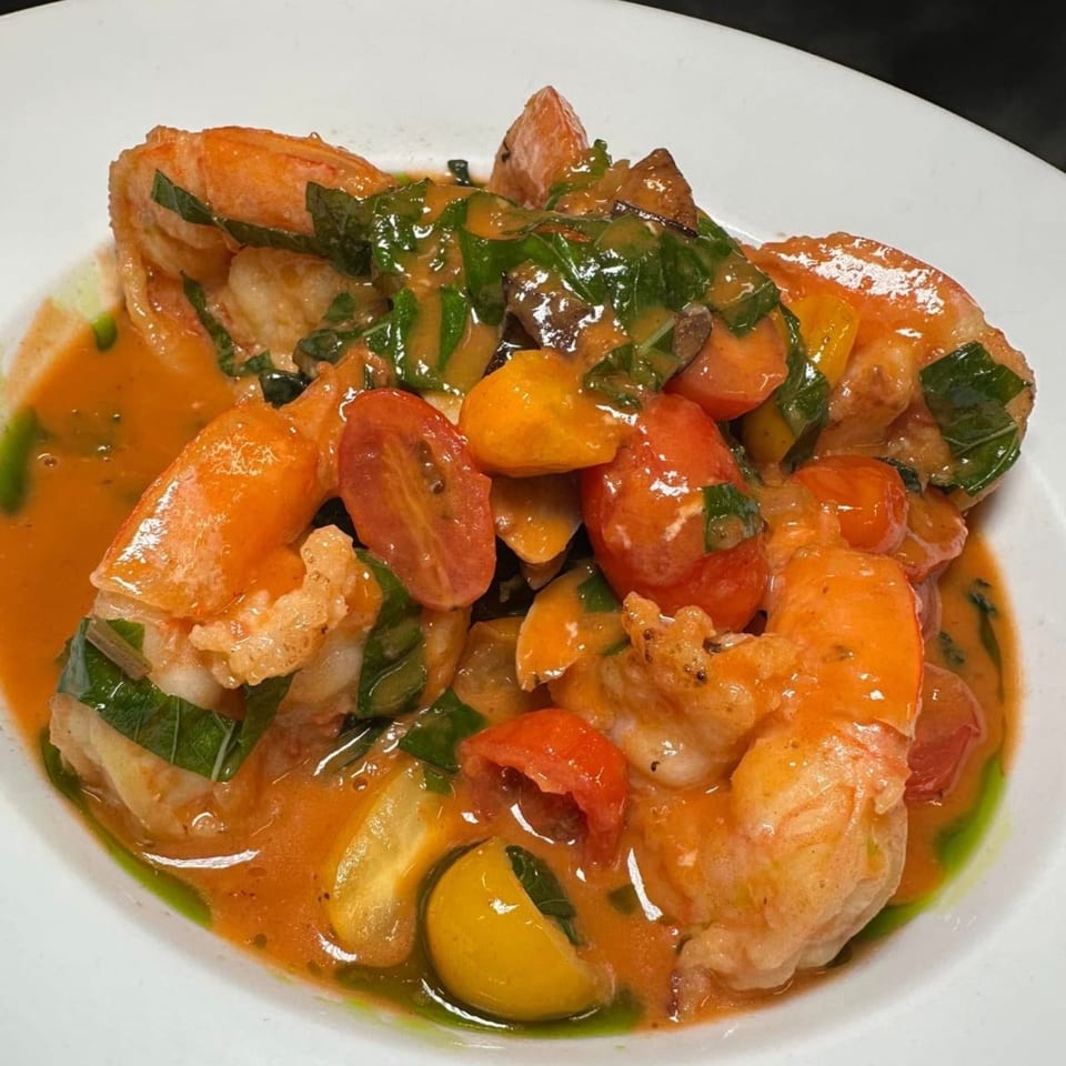 Shrimp in a rich orange sauce with halved cherry tomatoes and green herbs in a white bowl.